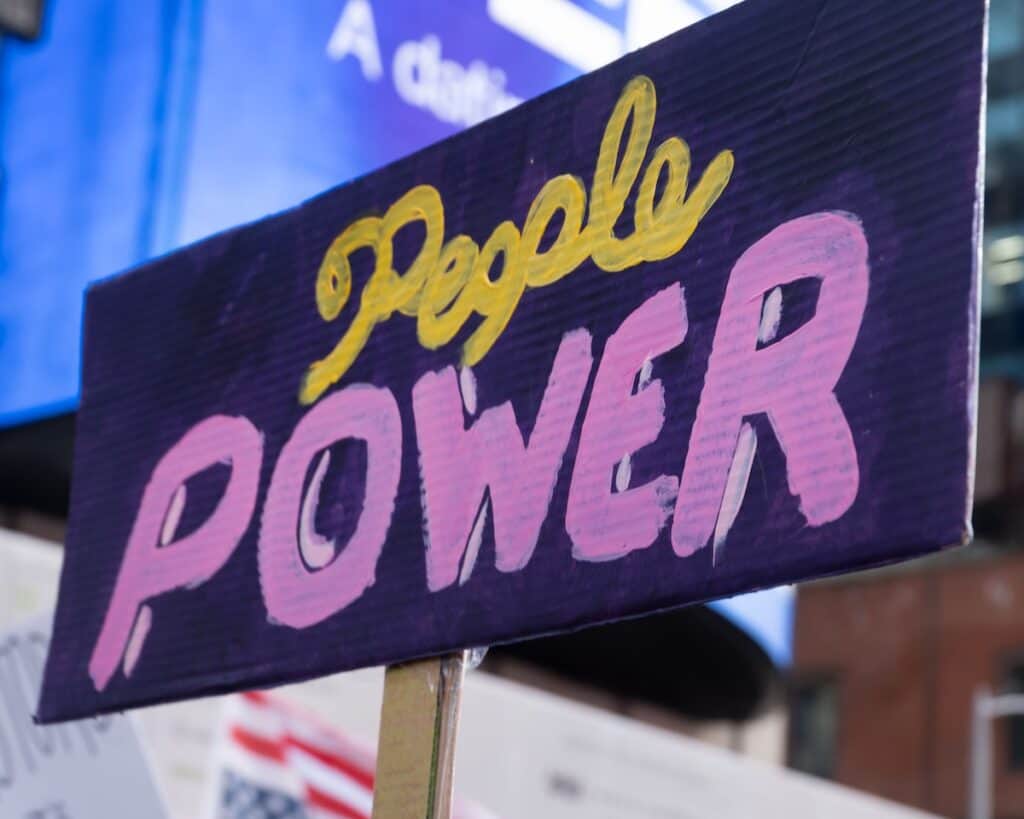 A colorful handpainted sign reads "People Power". Its maker used various shades of purple as well as yellow. Part of a giant display screen adds a lighter blue background. This photograph was made during the No Kings protest march, in the Borough of Manhattan, New York City, on October 18, 2025.