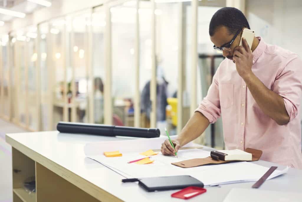 Black owner of building company solving issues via telephone conversation with business partner about architectural project standing at desktop with sketches near advertising area