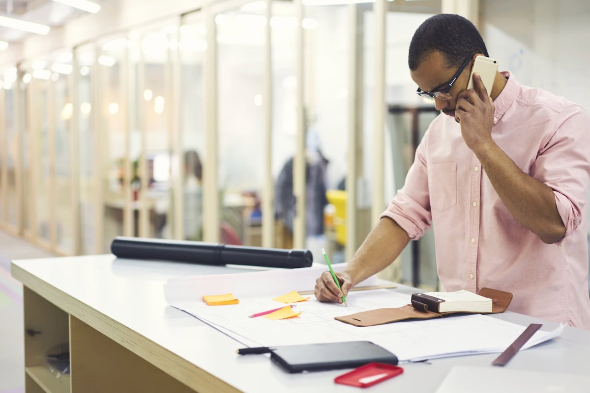 Black owner of building company solving issues via telephone conversation with business partner about architectural project standing at desktop with sketches near advertising area